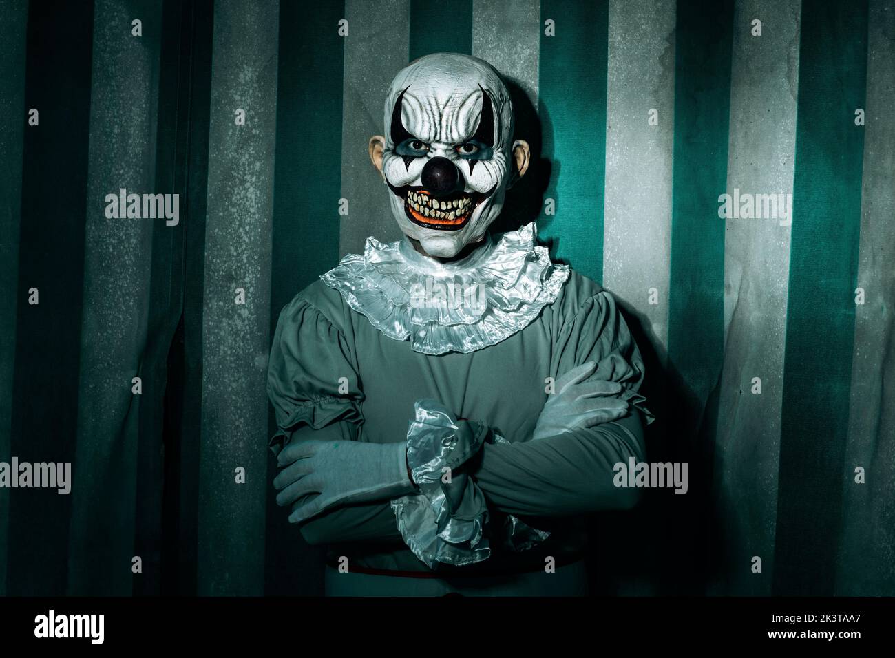 closeup of a creepy bald evil clown, standing in front of a dirty and old circus tent, wearing a gray costume with a white ruff, and starring at the o Stock Photo