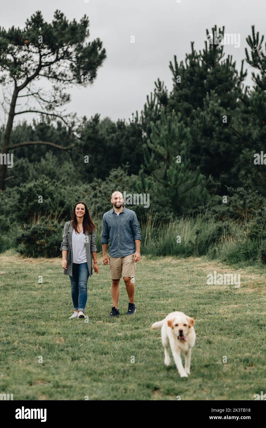 Happy couple in casual wear walking with dog in green park Stock Photo ...