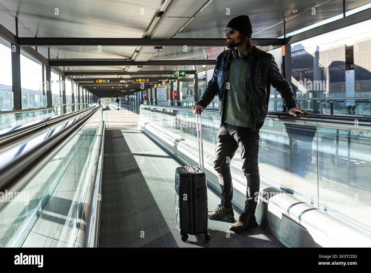 Hipster man in black leather jacket and knitted hat walking with suitcase on treadmill in airport terminal Stock Photo
