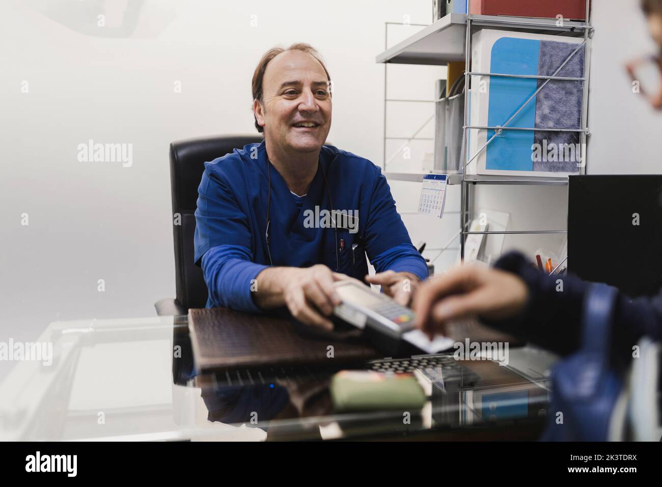 Positive adult male doctor in uniform giving payment terminal with