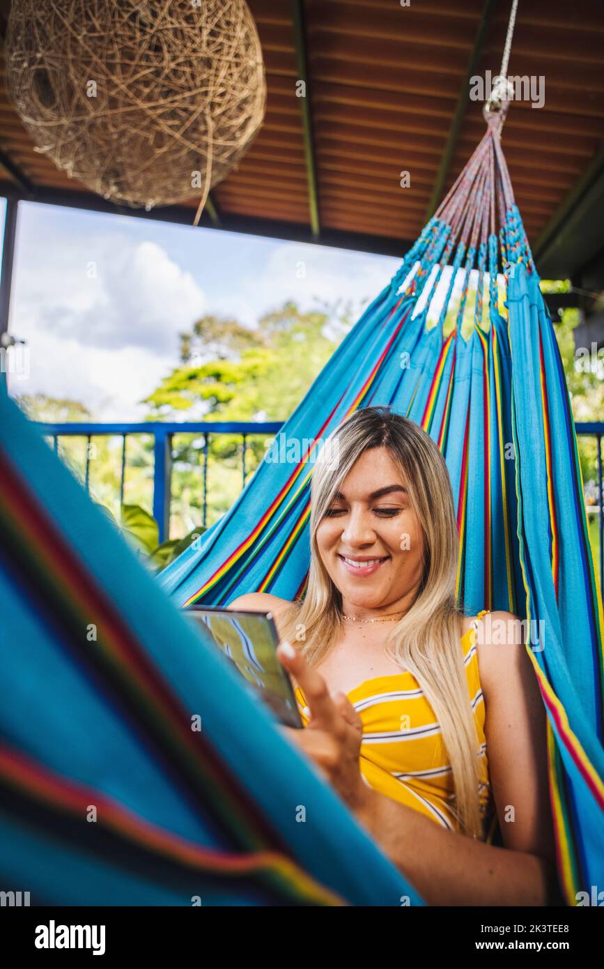 Cheerful young female in yellow sundress using smartphone and chilling in hammock on resort terrace Stock Photo