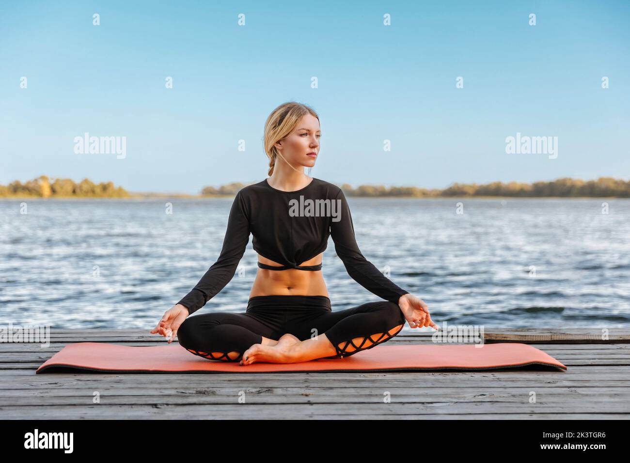 A young beautiful girl is doing yoga in a calm warm evening on the banks of the river. Stock Photo
