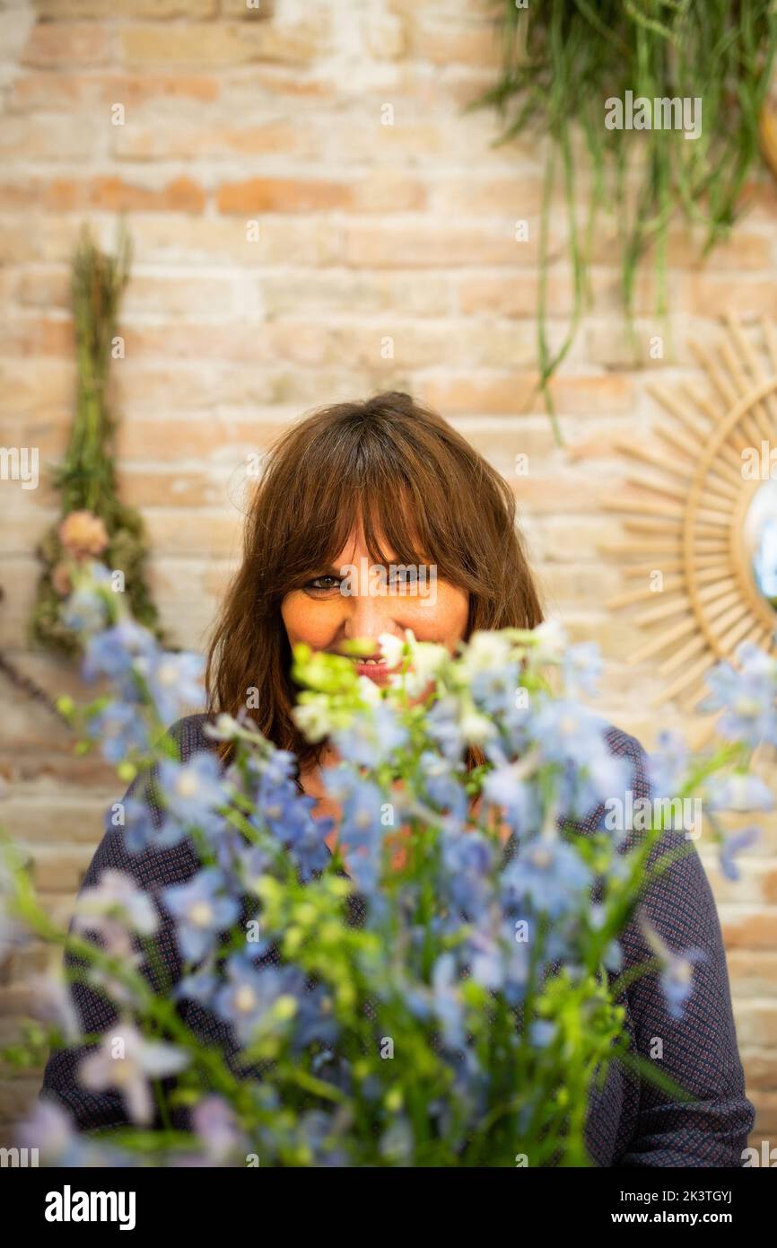 Smiling woman touching and adjusting delicate blue flowers in fresh bouquet during work in flower store looking at camera Stock Photo