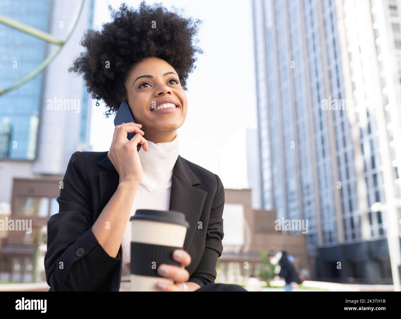 Smiling African American female architect in formal suit sitting on street with takeaway coffee and talking on smartphone while discussing project and Stock Photo