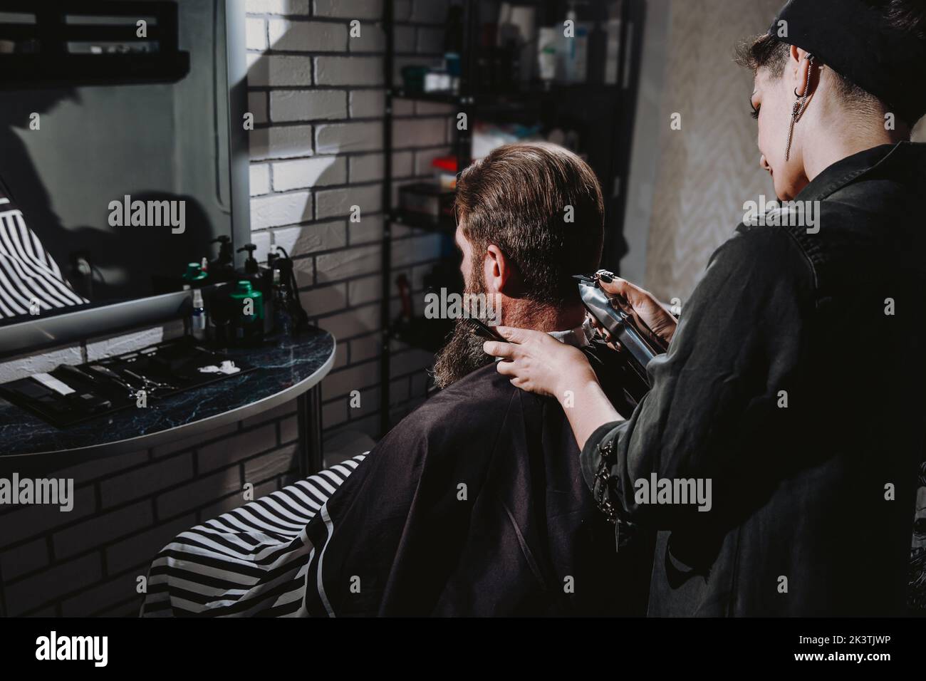 Young woman barber making haircut of bearded man in barbershop. Self-care, masculine beauty. Stock Photo