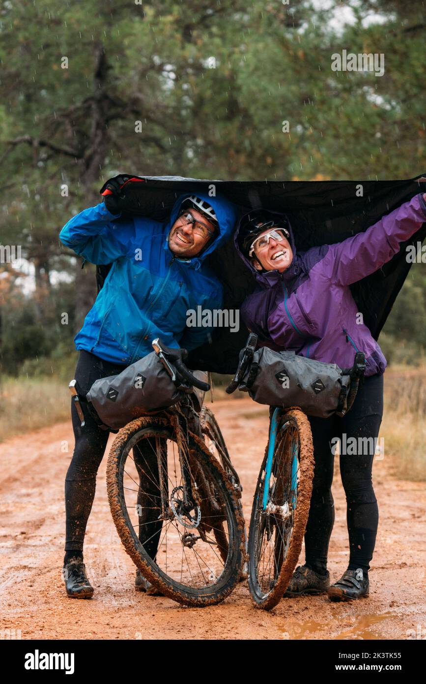 Active well equipped people in protective helmet and glasses leaning on bikes and having fun with tent pulling over heads on dirty road in forest Stock Photo