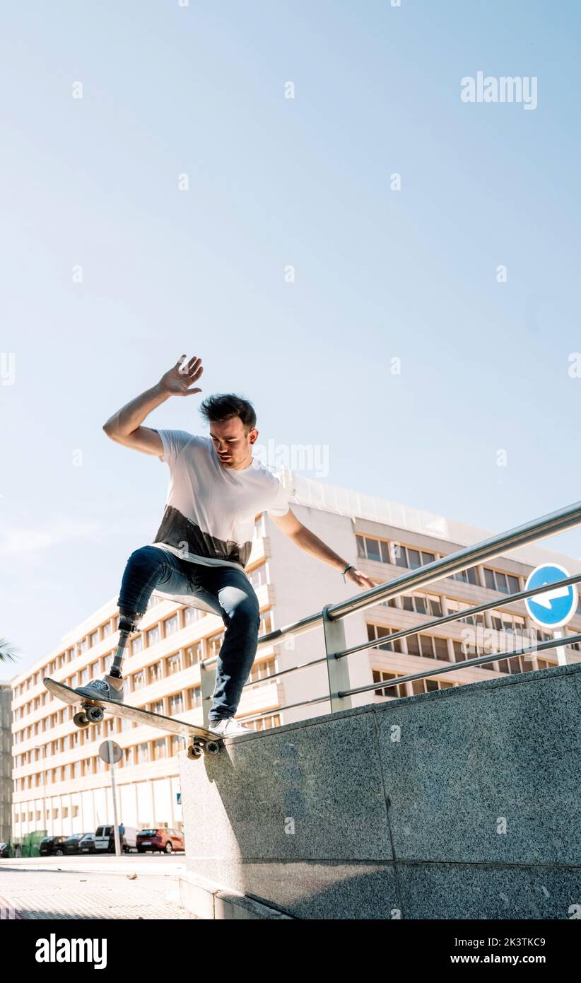 From below young guy with leg prosthesis jumping on skateboard above ground against modern building on sunny day Stock Photo