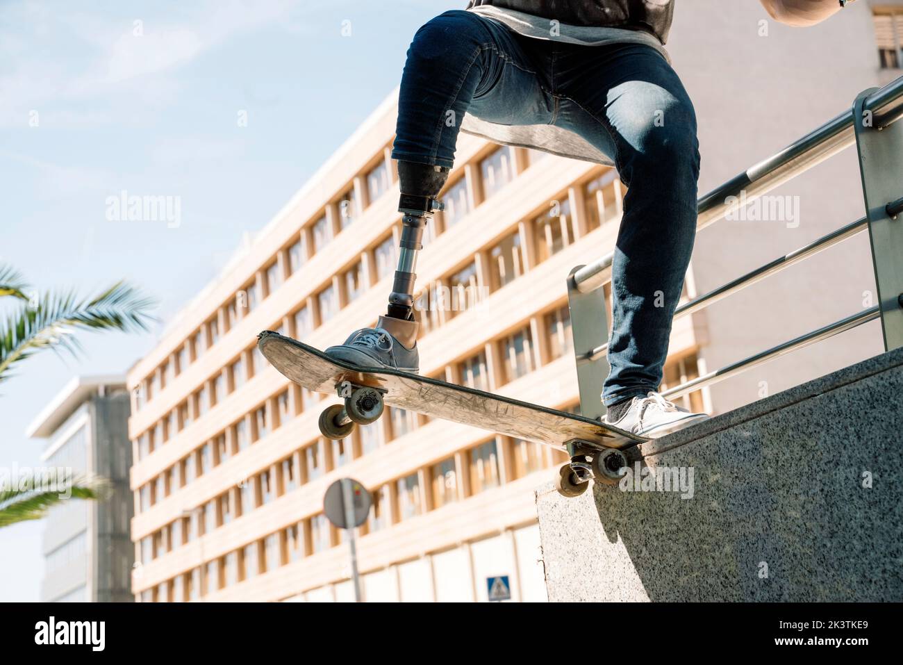 From below young guy with leg prosthesis jumping on skateboard above ground against modern building on sunny day Stock Photo