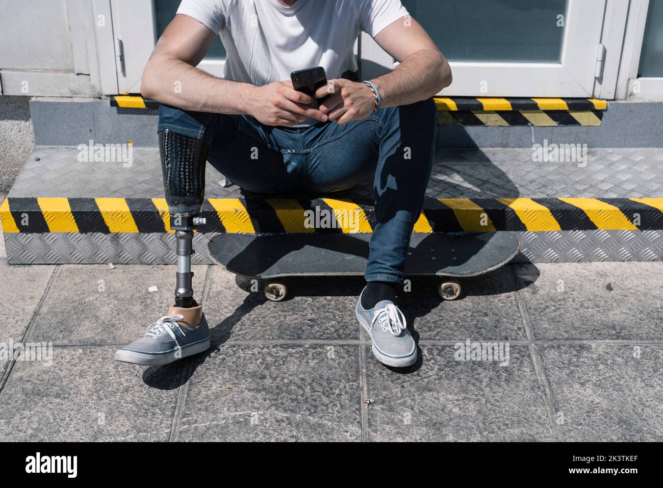 From above anonymous male with artificial leg browsing smartphone while sitting on step near skateboard on sunny day on city street Stock Photo