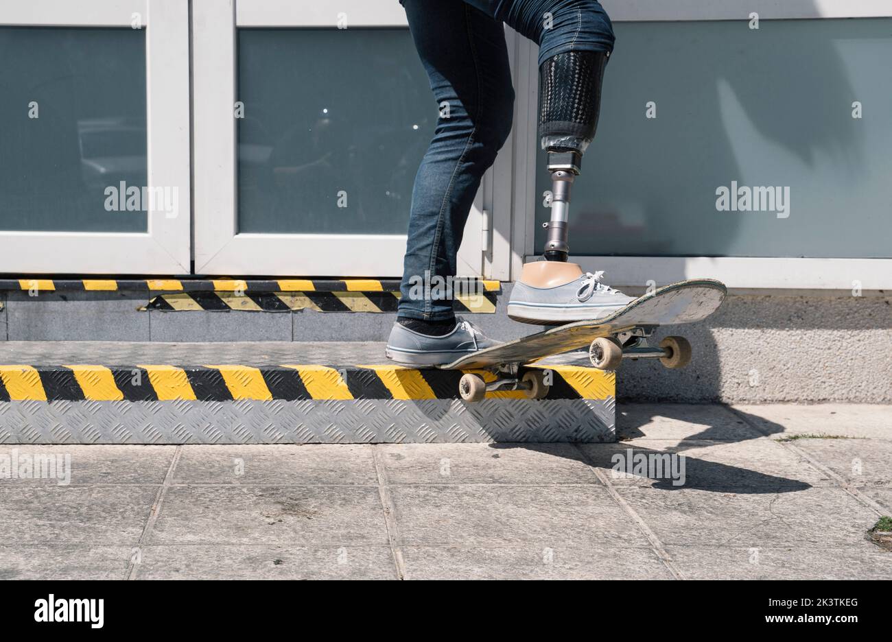 From below young guy with leg prosthesis jumping on skateboard above ground against modern building on sunny day Stock Photo