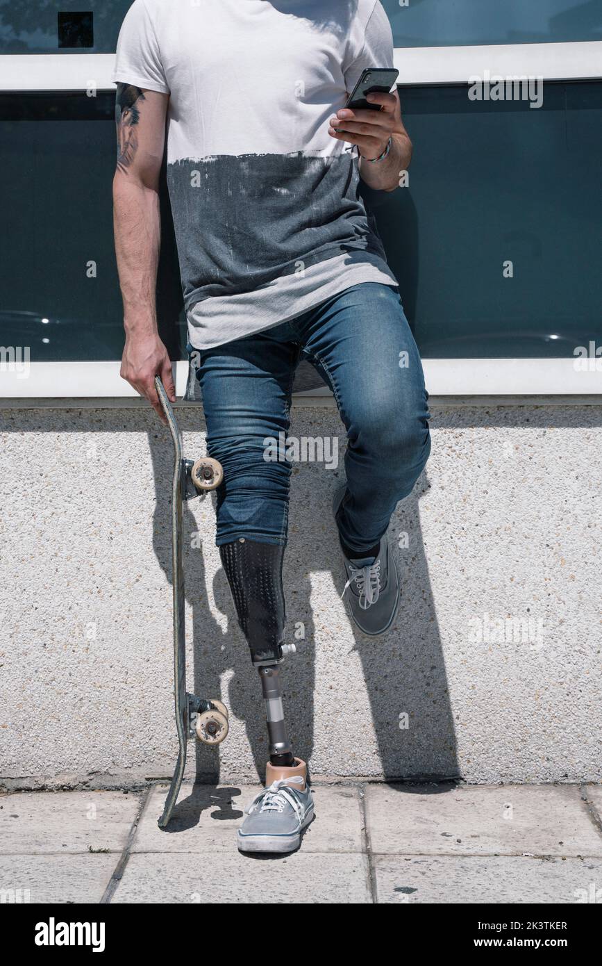 From above anonymous male with artificial leg browsing smartphone while standing holding skateboard on sunny day on city street Stock Photo
