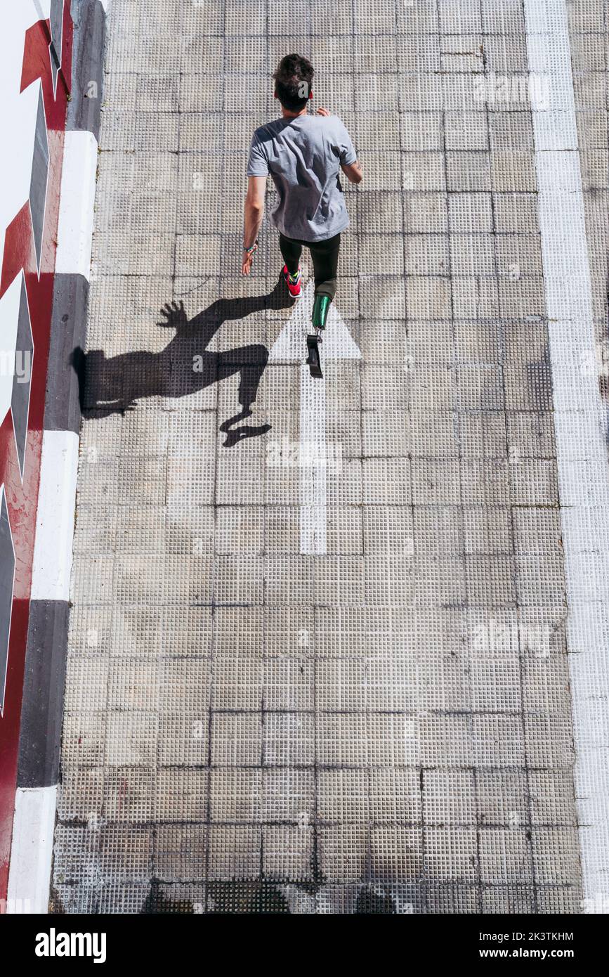 from above young guy with leg prosthesis running up paved slope against shabby building during workout on city street Stock Photo