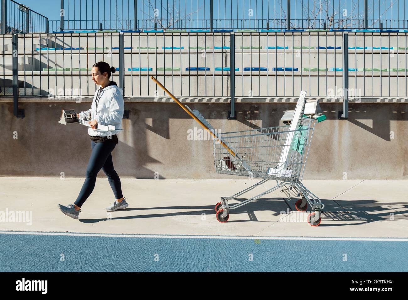 Side view of young female athlete carrying starting blocks while walking near cart with gear before track and field training on sunny day on stadium Stock Photo