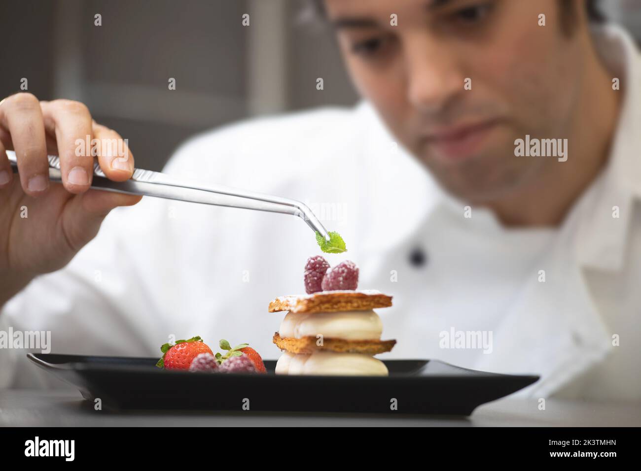 Crop focused man in white chef jacket decorating fancy dish with mint and raspberry on plate Stock Photo