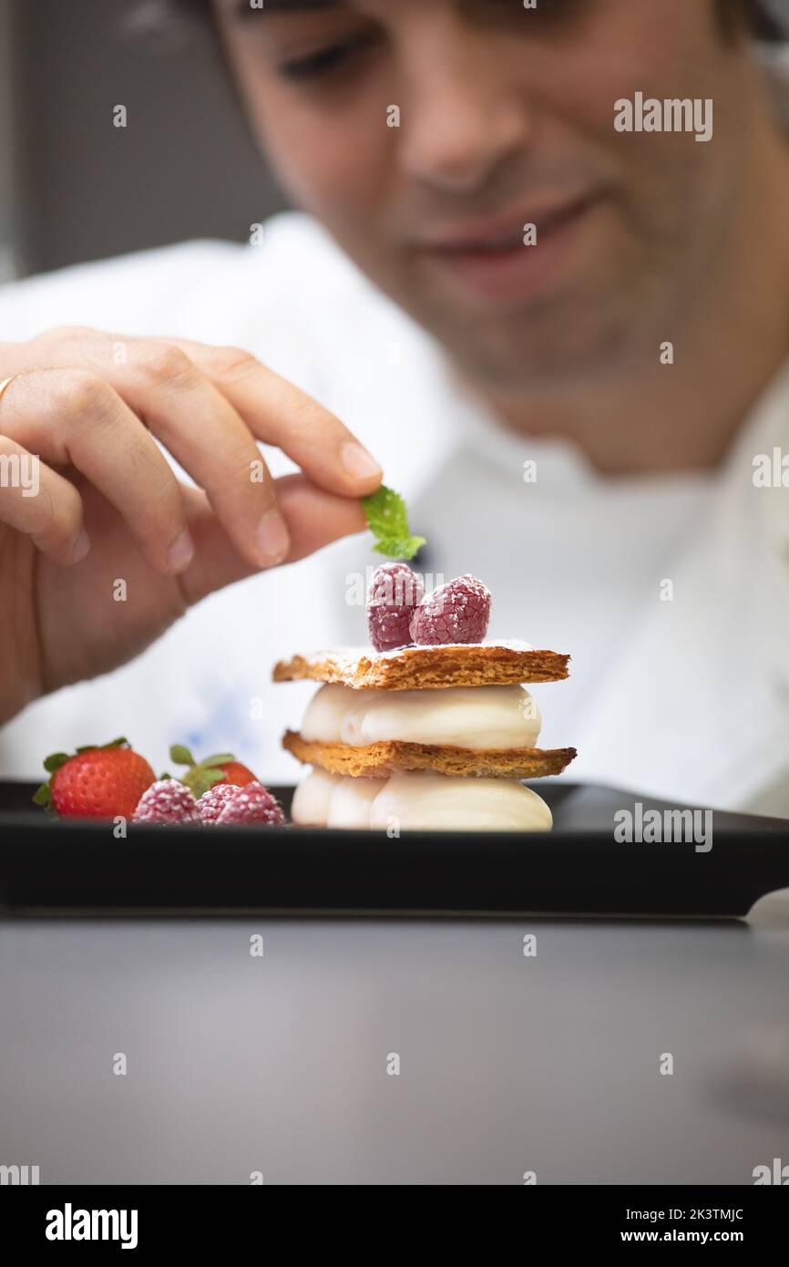 Crop focused man in white chef jacket decorating fancy dish with mint and raspberry on plate Stock Photo