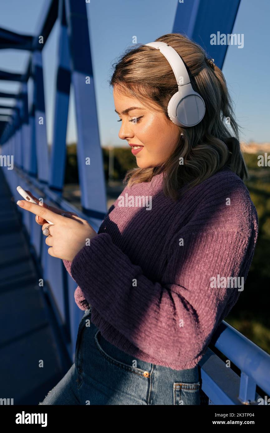 Low angle of cheerful female in headphones standing on bridge while listening to music and surfing Internet on smartphone Stock Photo