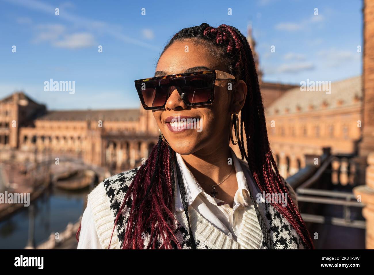 Cheerful African American female with braids standing on Plaza de Espana on sunny day and looking away during weekend in Seville Stock Photo