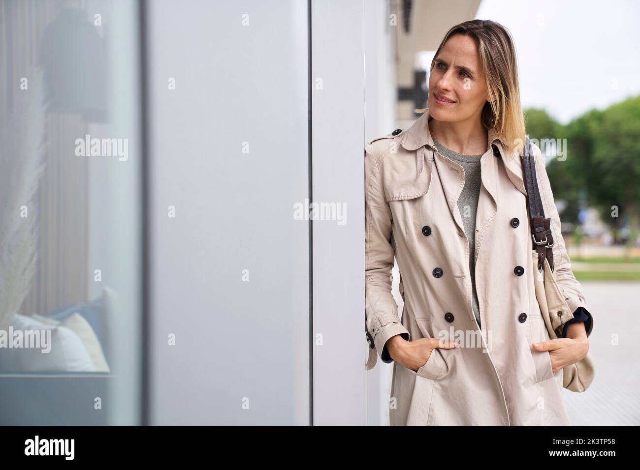 Mid-shot of woman wearing raincoat looking at shop window while leaning on a wall Stock Photo