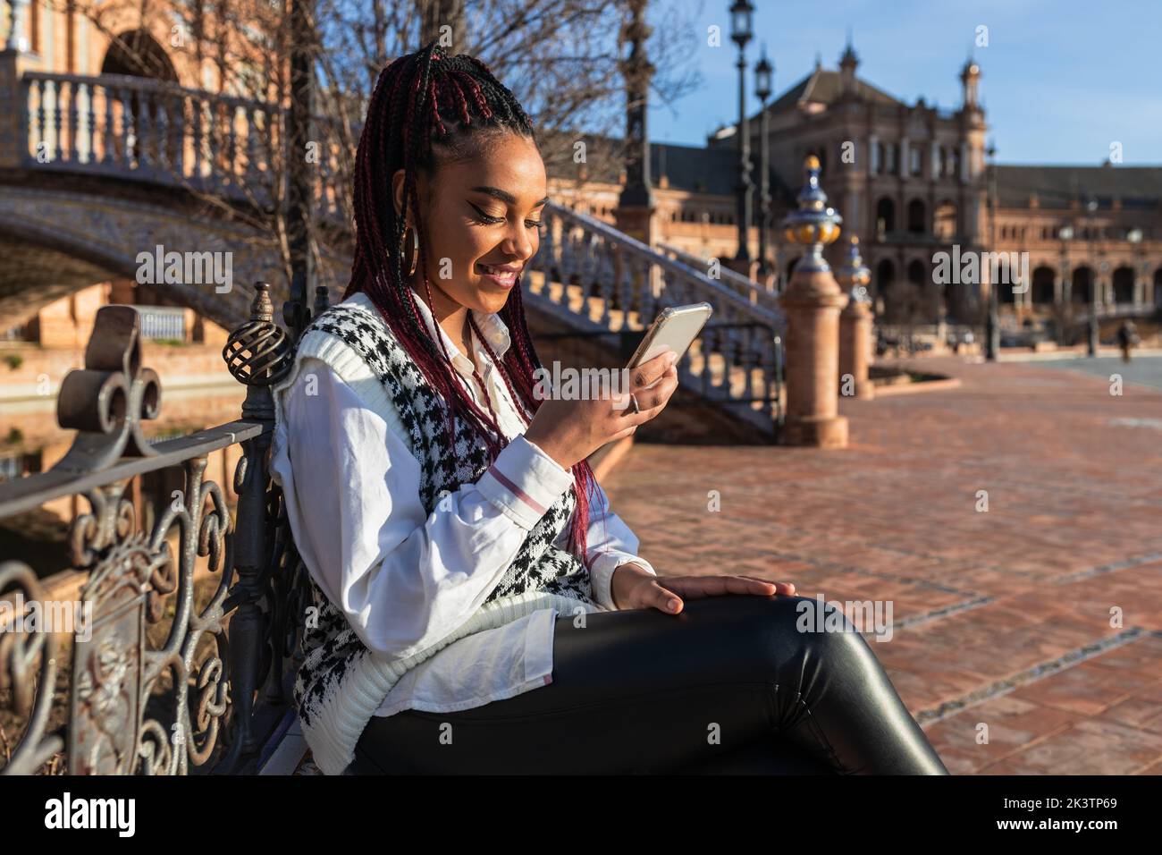 Side view of delighted African American female with braids and in stylish wear sitting on Plaza de Espana on sunny day and chatting on social media vi Stock Photo