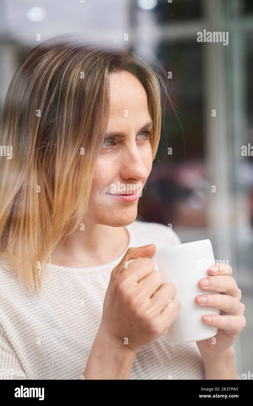 Mid-shot portrait of woman holding a coffee mug while looking through a window Stock Photo