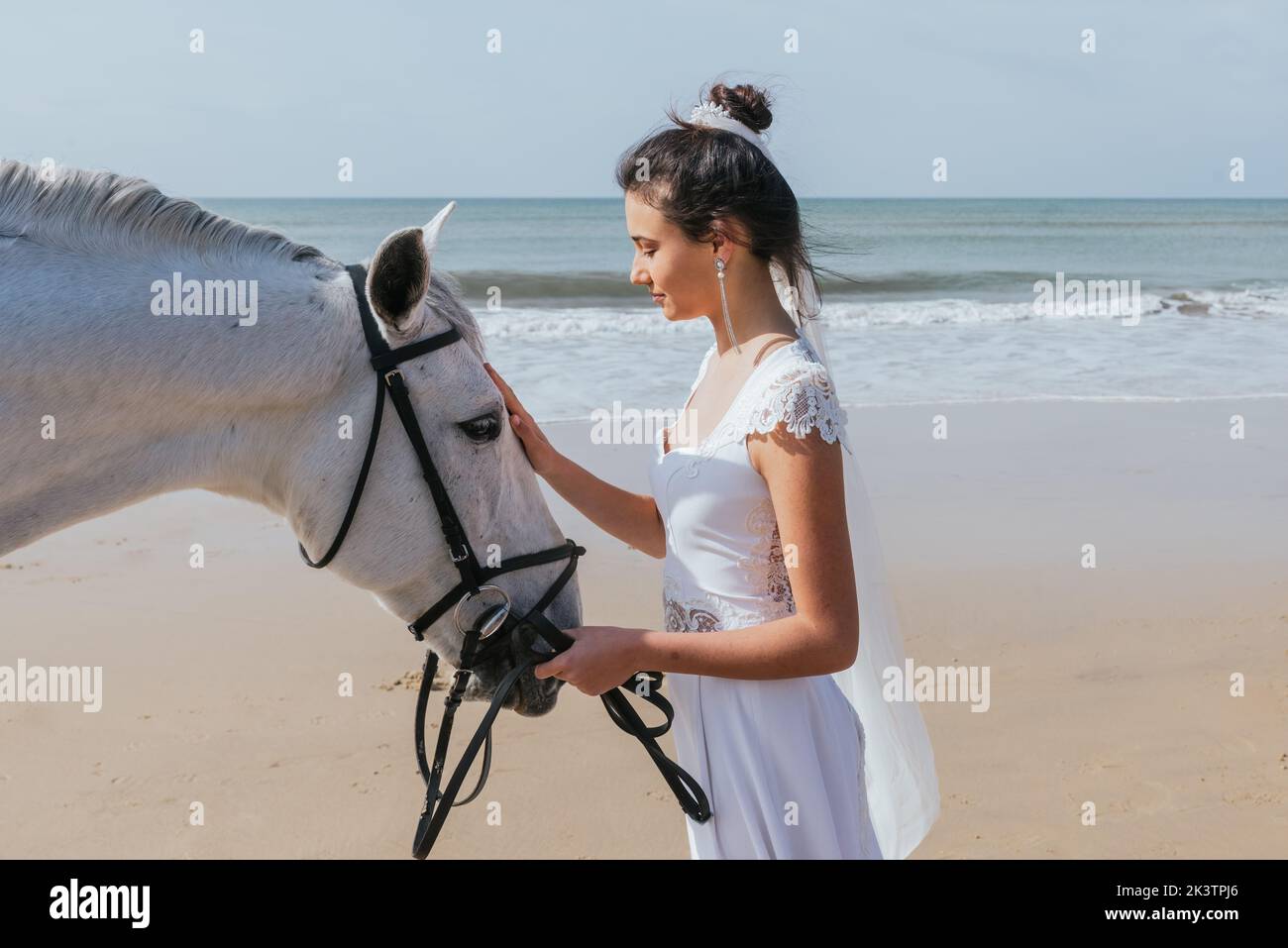 Side view of serene woman in white dress standing with purebred mare with reins along sandy ocean shore Stock Photo