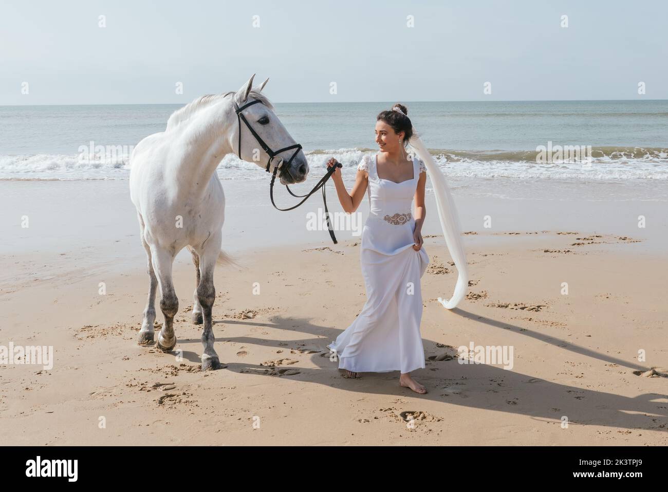 Cheerful woman in white dress leading purebred mare by reins while looking at each other on sandy ocean shore Stock Photo