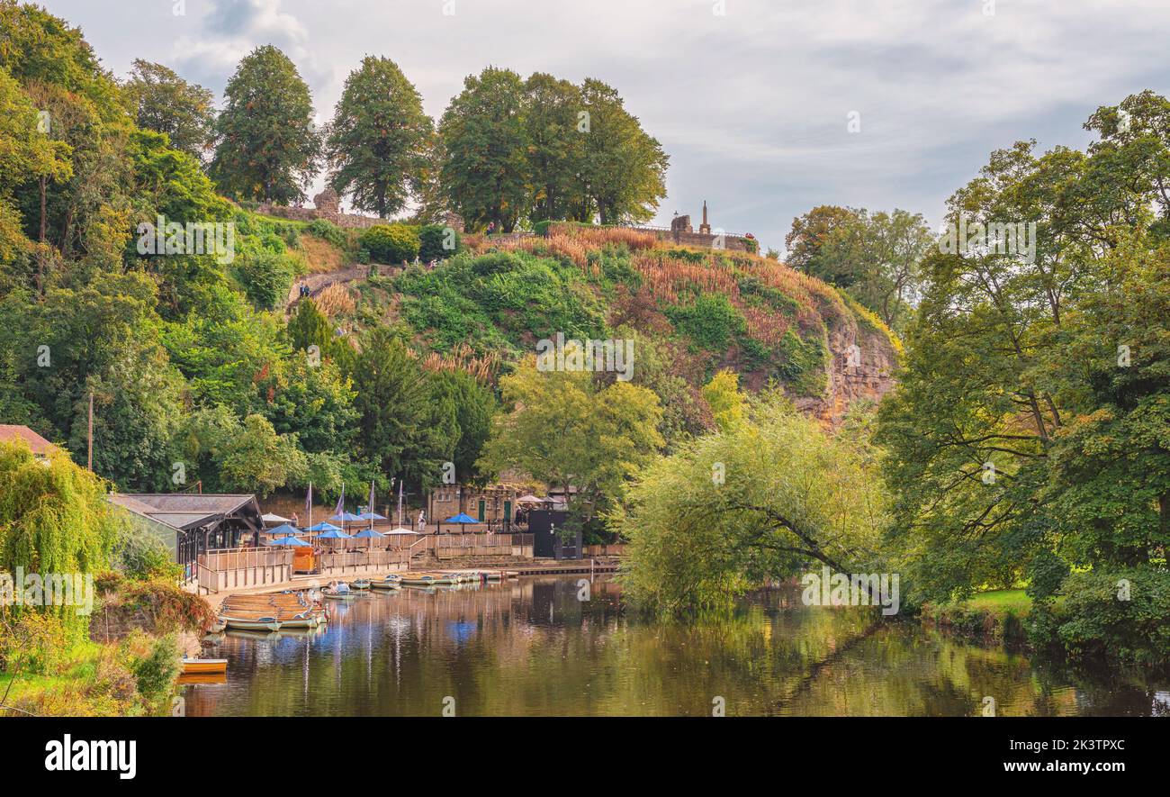 A tranquil scene where a tree arches over a river adjacent to moored boats. A hill is beyond with a monument on the skyline. A cloudy sky is above. Stock Photo