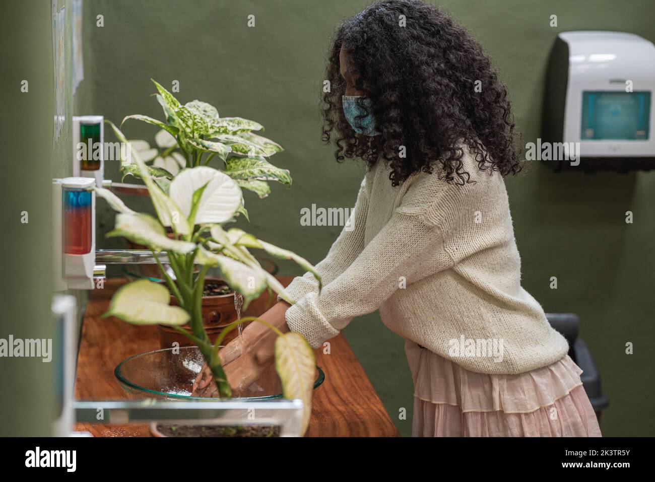 Side view of ethnic female in disposable mask washing hands in washbasin in house with tropical plants Stock Photo