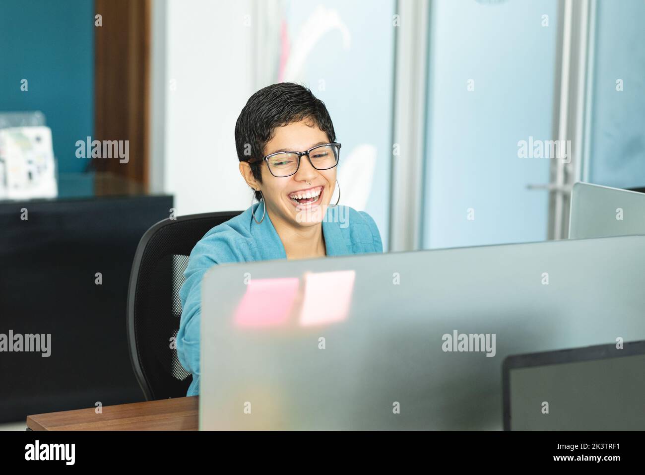 Cheerful Hispanic female entrepreneur with short hair in stylish outfit sitting at table with laptop and laughing while working in light office in Cos Stock Photo