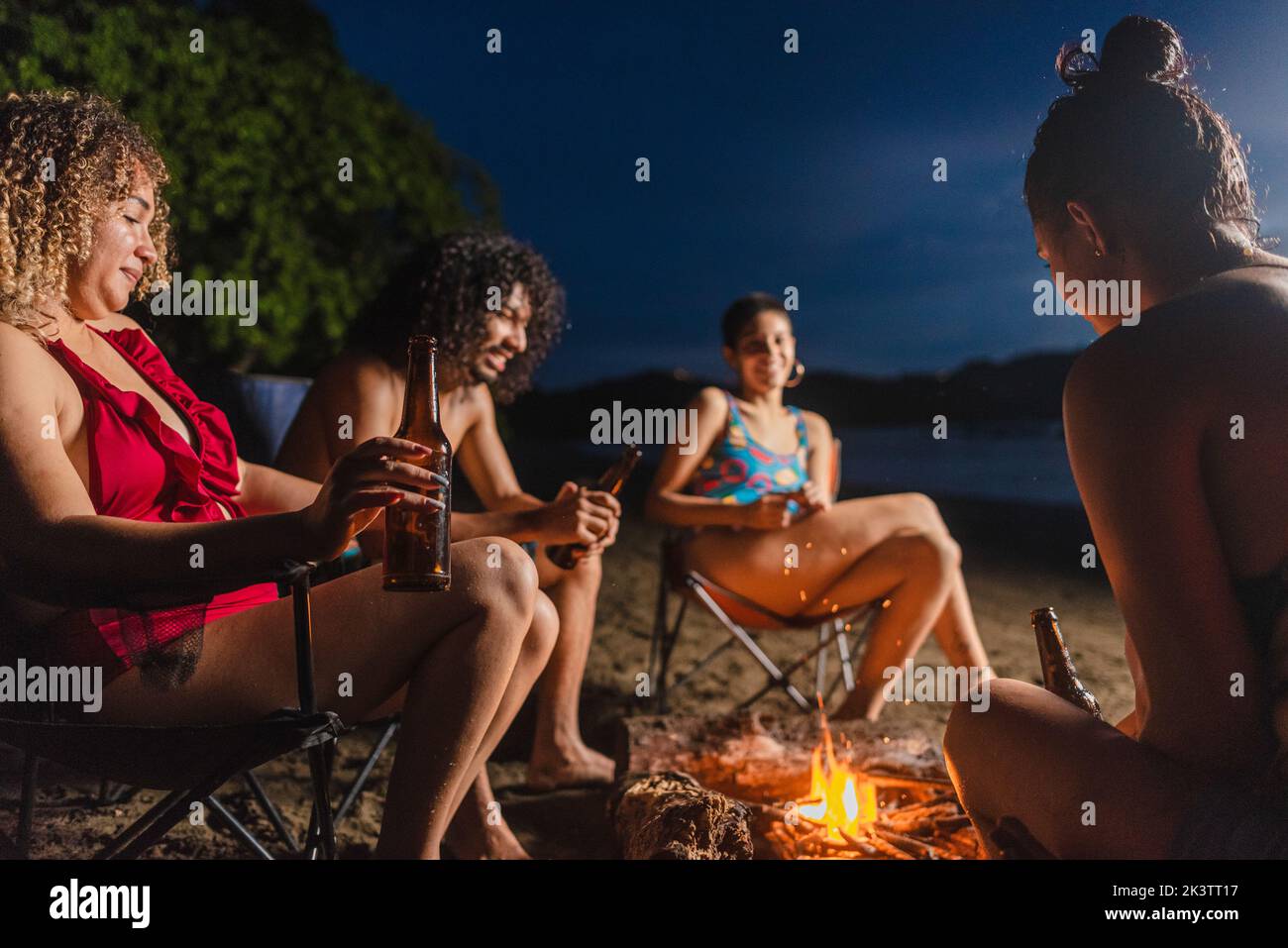Group of cheerful ethnic friends with beer having fun while gathering around burning bonfire during summer hangout in evening time on beach in Costa R Stock Photo