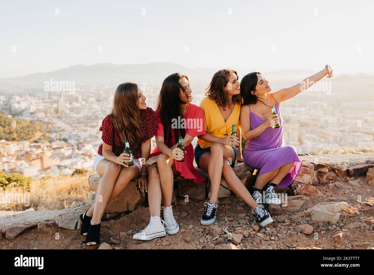 Cheerful group of young female friends in casual outfits taking selfie on smartphone while sitting on rocky ground on hilltop and drinking beer Stock Photo