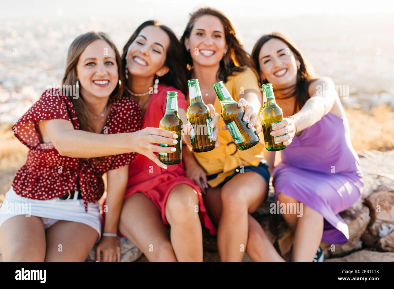 Group of joyful female friends toasting to the camera with glass bottles of beer while siting on hilltop over town against cloudless sky in sunlight Stock Photo