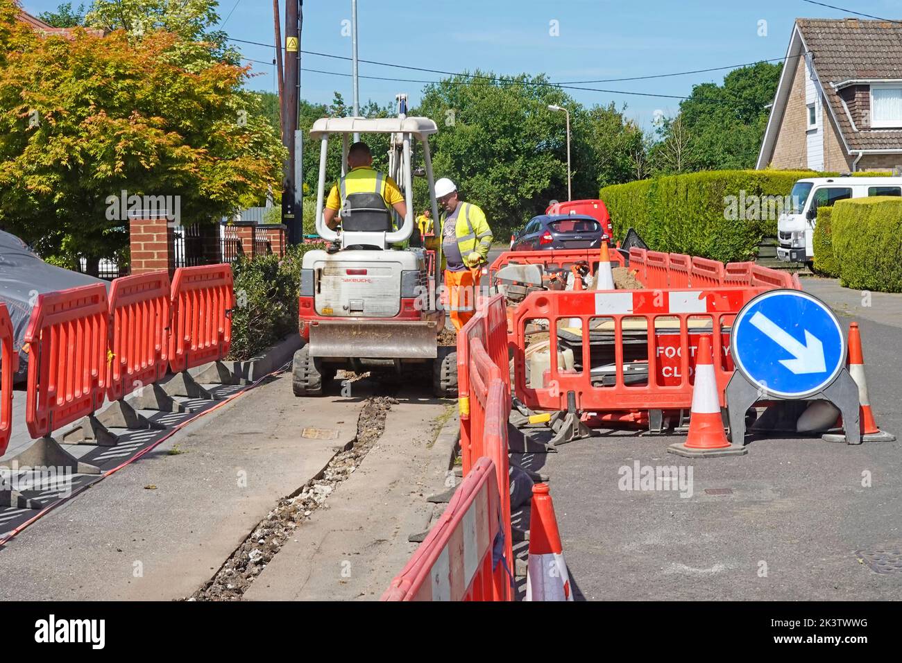 Red road works plastic safety barriers restricting home owners driveway access mini excavator digging broadband infrastructure trench in pavement UK Stock Photo