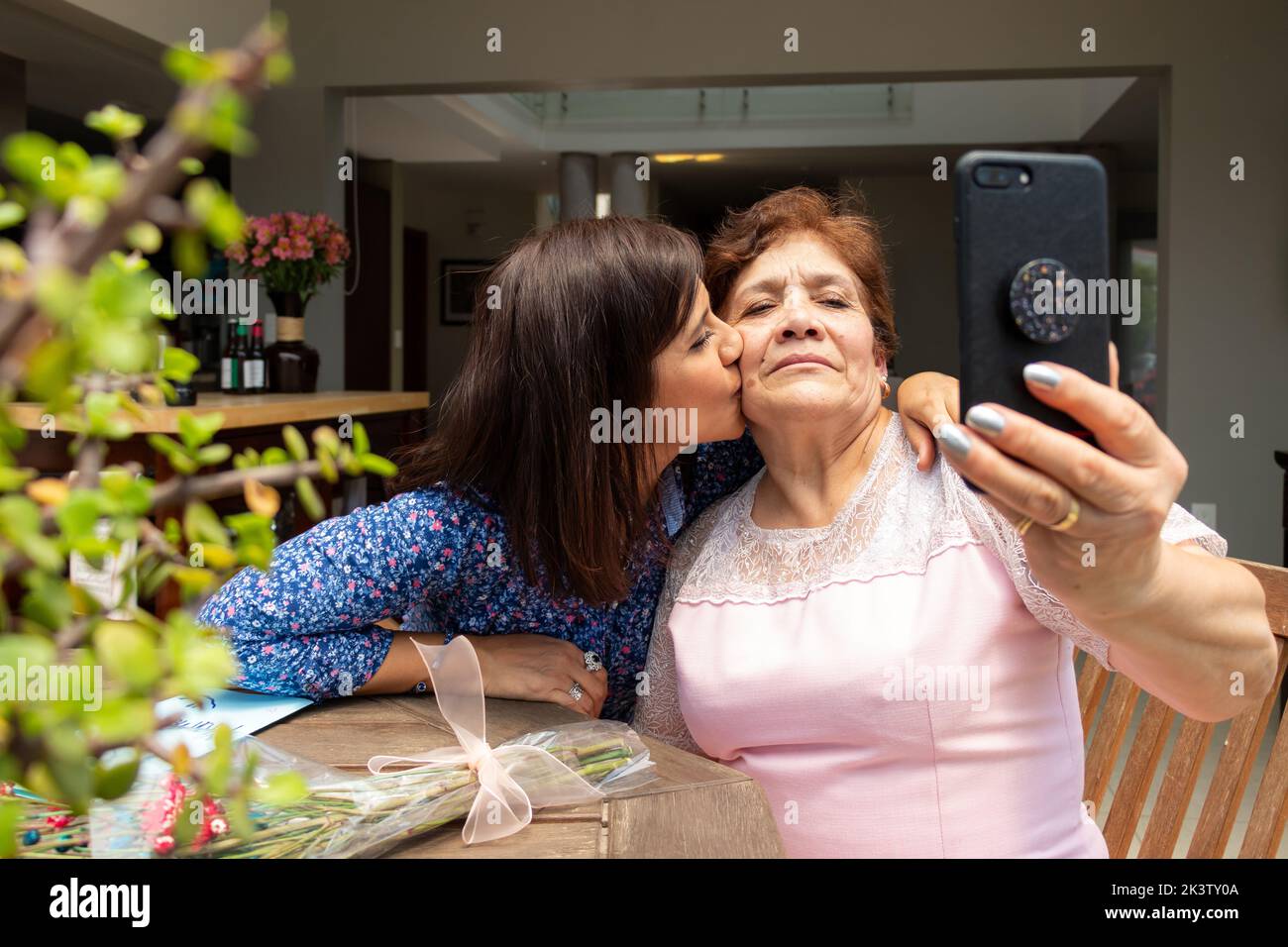 Happy ethnic adult woman kissing cheek of content senior mother taking selfie on mobile phone sitting at table on terrace Stock Photo
