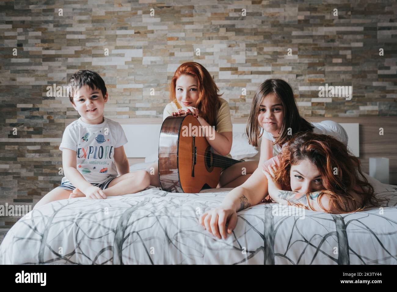 Young same gender parents in pajamas playing guitar with kids and looking at camera on bed in bedroom Stock Photo