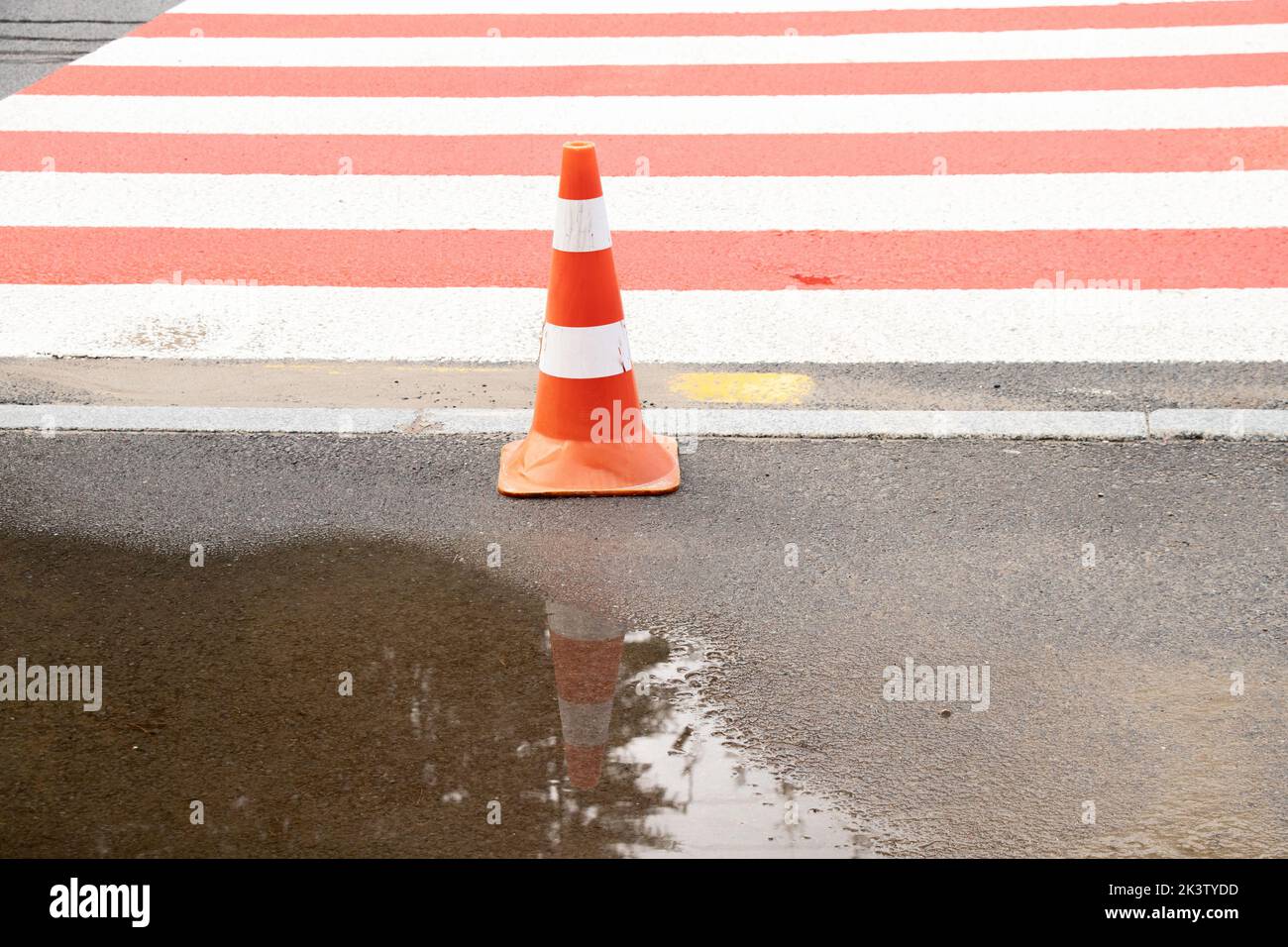 A striped road cone stands on a zebra crossing near a puddle in autumn in Ukraine Stock Photo