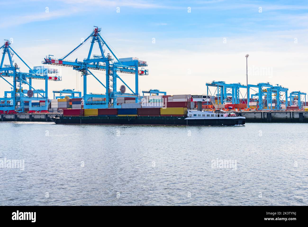 Cargo barge loaded with colourful containers sailing past a comercial dock with tall gantry cranes and stacks of containers Stock Photo