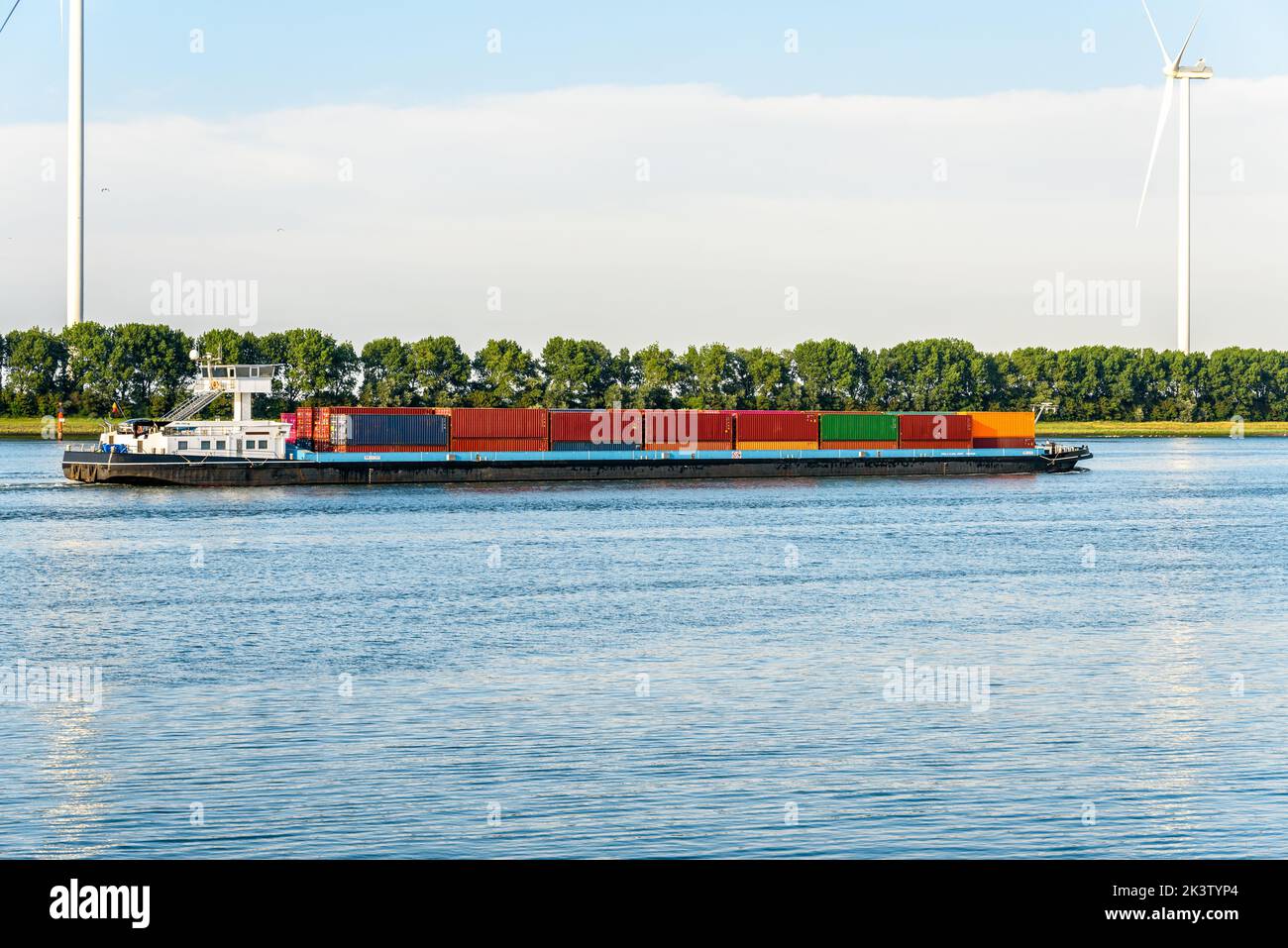 Cargo barge loaded with containers sailing up a river at sunset. Wind turbine are on the riverbank. Stock Photo