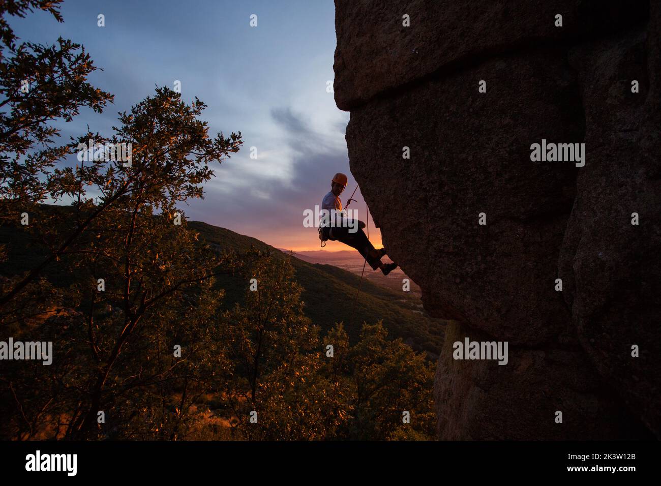 Side view of active climber in protective helmet hanging on mountain holding on to slope in warm light of sunset Stock Photo
