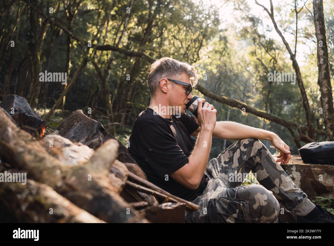 Side view of mature male traveler in sunglasses drinking hot beverage from thermos cup while sitting in campsite Stock Photo