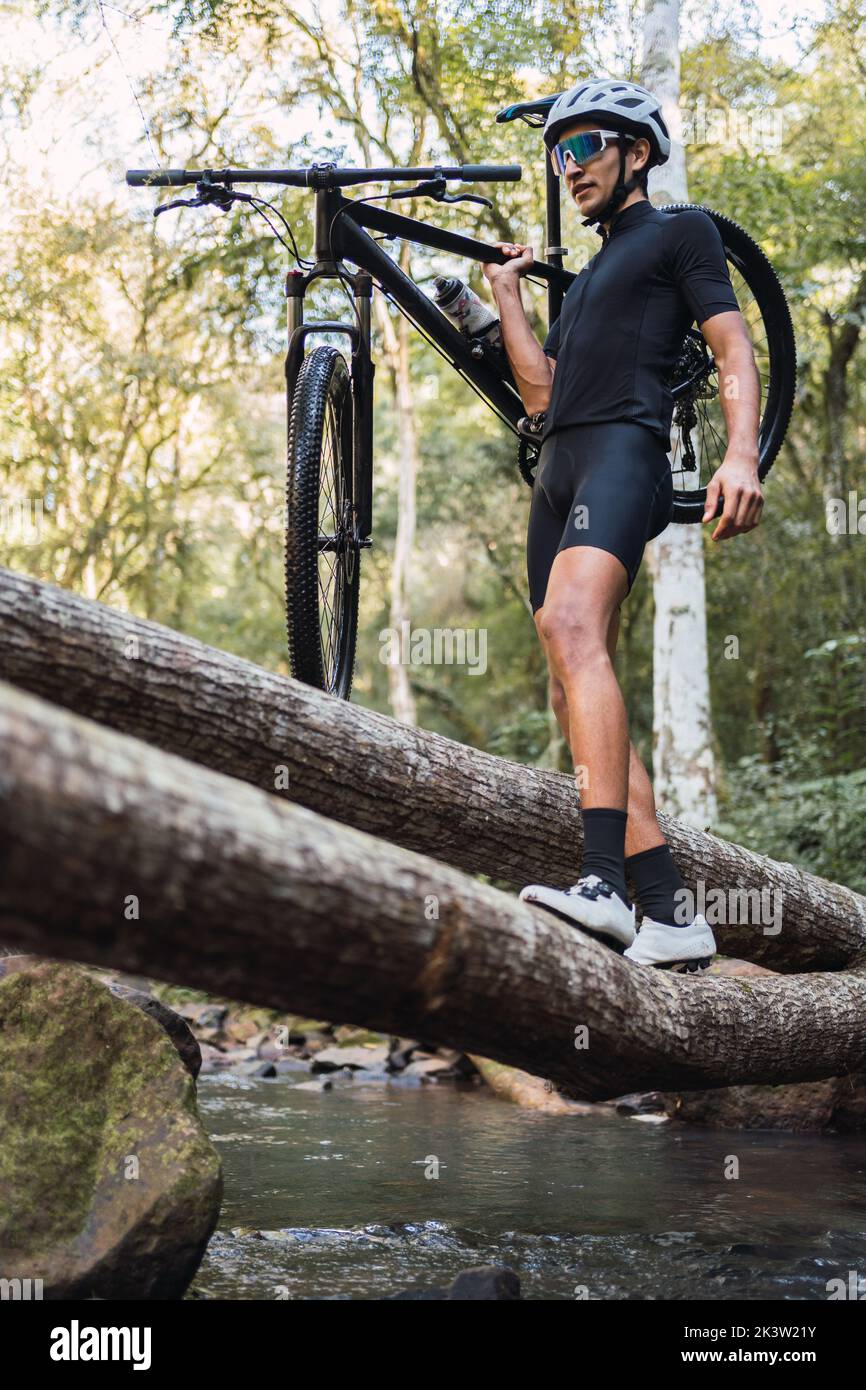Low angle of fit sportsman in helmet and cycling goggles carrying bicycle on shoulder while walking on fallen tree trunk in woods Stock Photo
