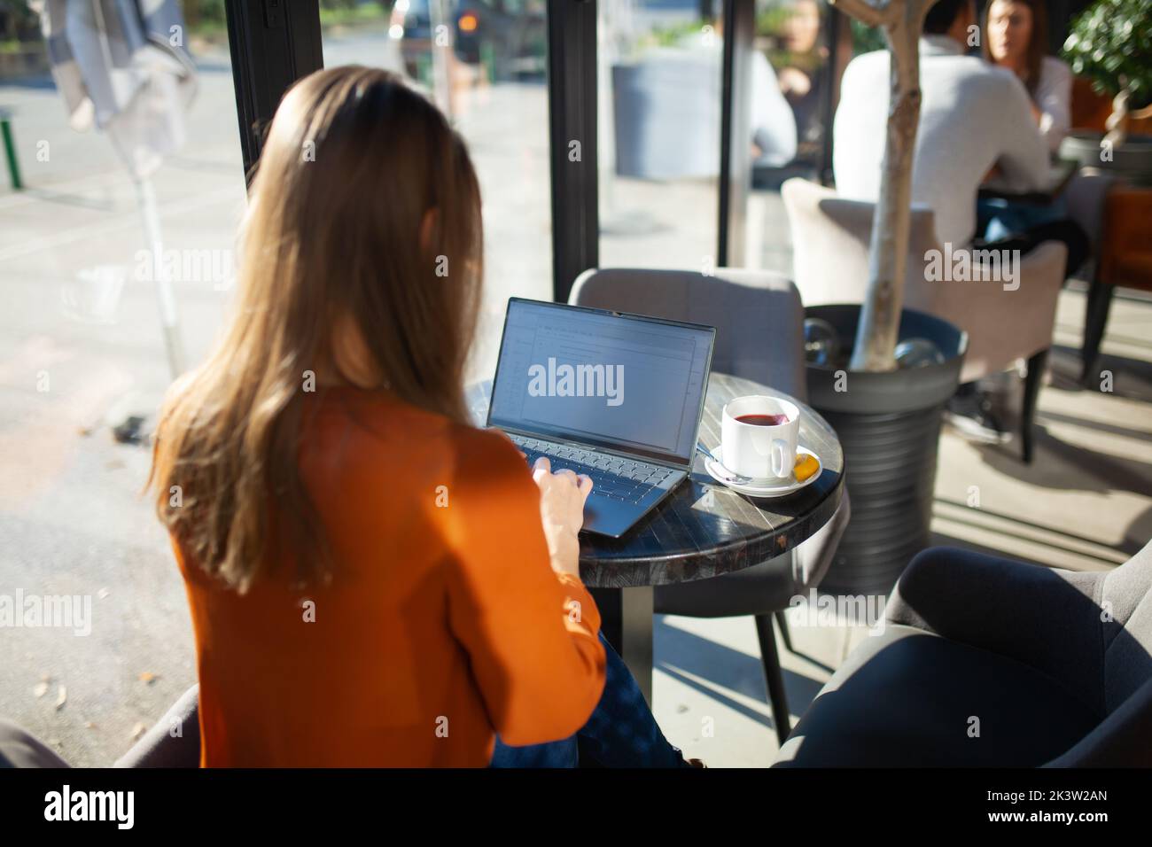 Young businesswoman in a cafe bar or restaurant with bright sunlight. Freelancer girl working on laptop and having tea at a window table. Stock Photo