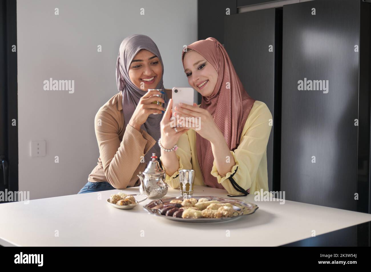 Two muslim women laugh and use the phone while having tea and arabic sweets. Stock Photo