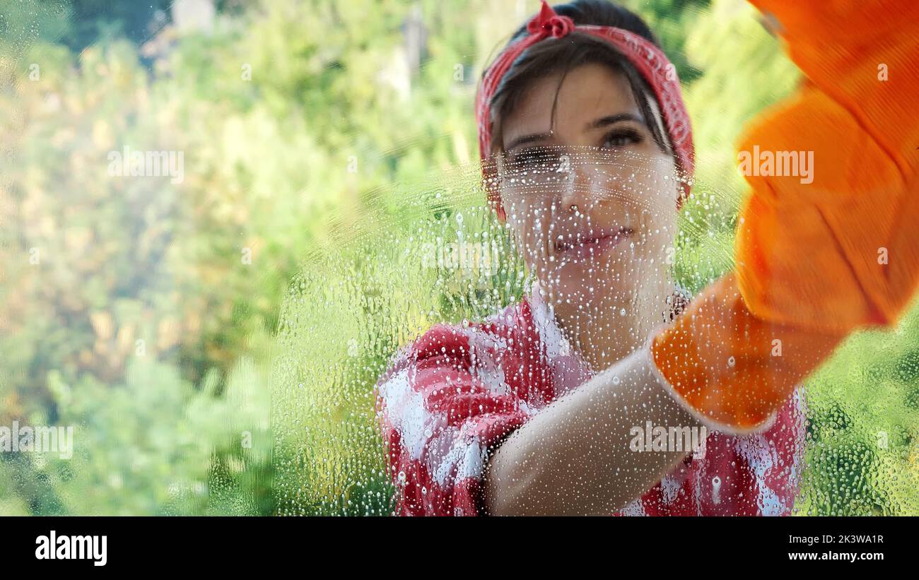 view through the window, smiling Beautiful woman in gloves, Cleaning Window by spraying Cleaning Products, using detergent and rag. worker of cleaning service or housewife. High quality photo Stock Photo