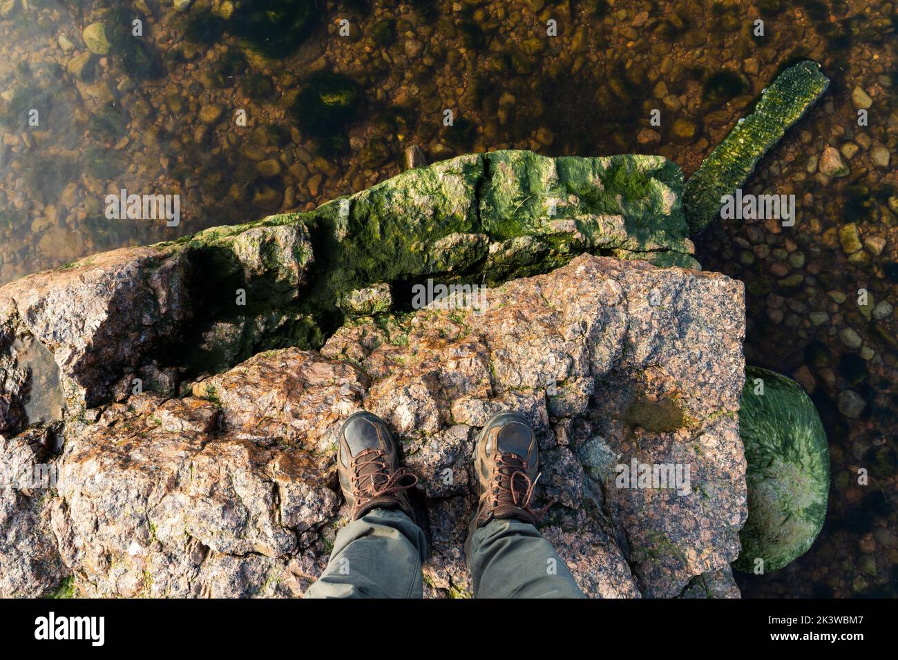 Feet of a man in rough trekking shoes standing on a coastal stone. Travel lifestyle vertical photo Stock Photo