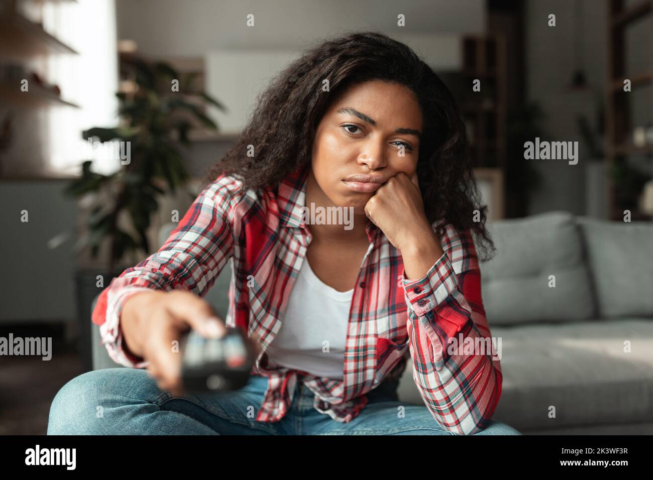 Bored sad pretty millennial african american female with remote control, watch tv news or movie, sitting on sofa Stock Photo