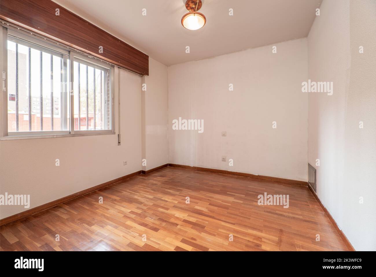 Empty room with reddish parquet flooring, plain white painted walls and aluminum window with bars Stock Photo
