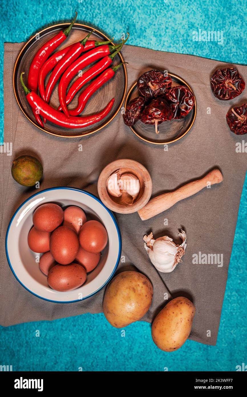 Still life with a plate of raw eggs, raw potatoes, garlic in a wooden mortar, dried peppers and red chili peppers on gray cloth and blue background Stock Photo