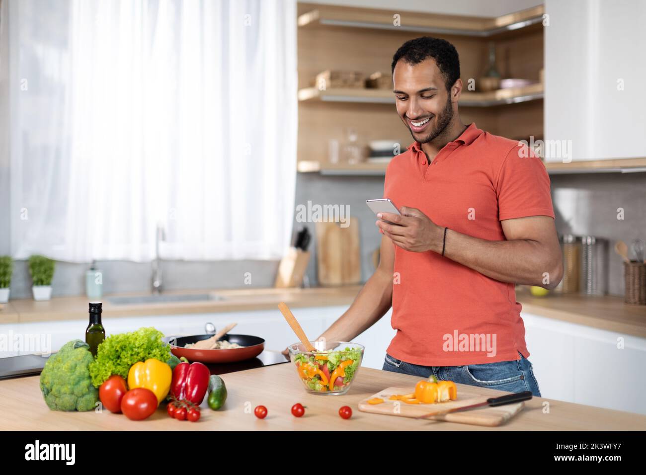 Cheerful young black male prepare salad at table with organic vegetables, chatting on smartphone Stock Photo