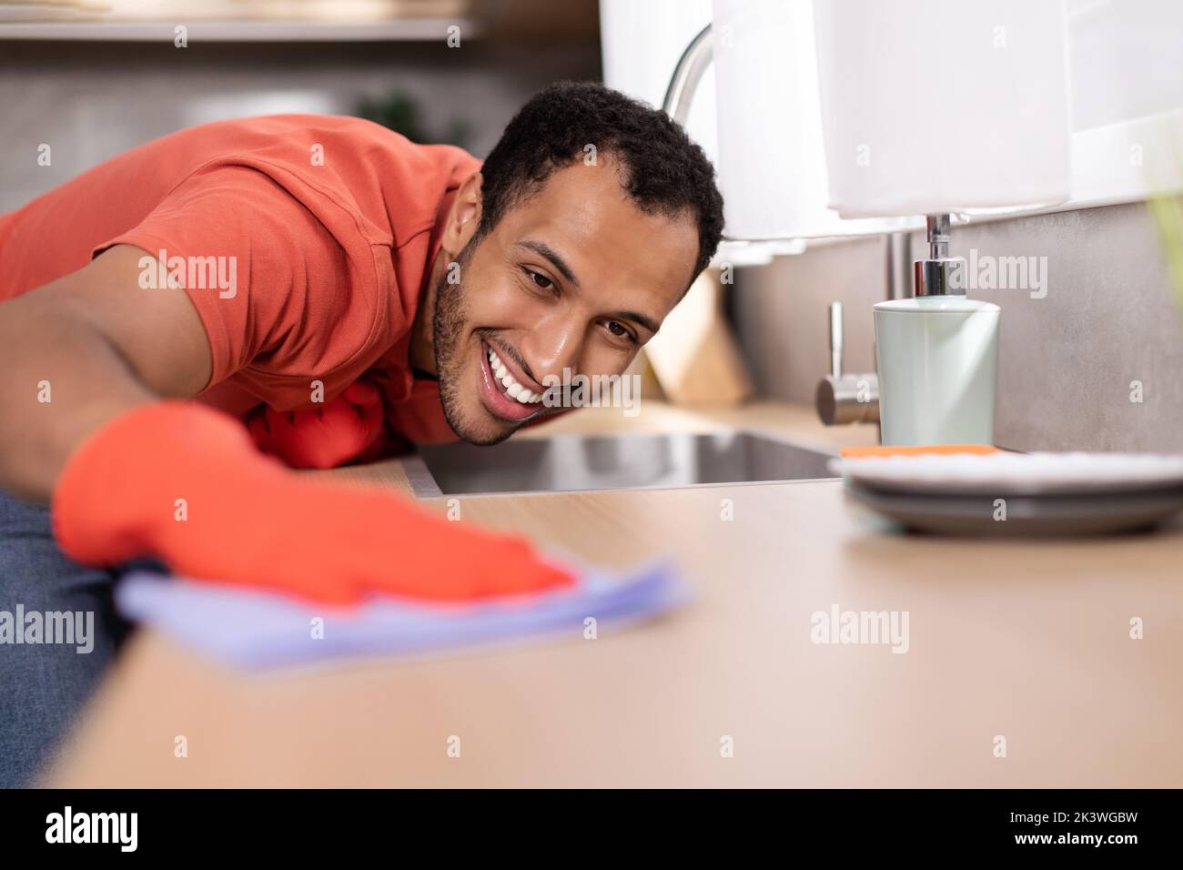 Smiling millennial black male in rubber gloves wipes dust from wooden table in kitchen interior, free space Stock Photo